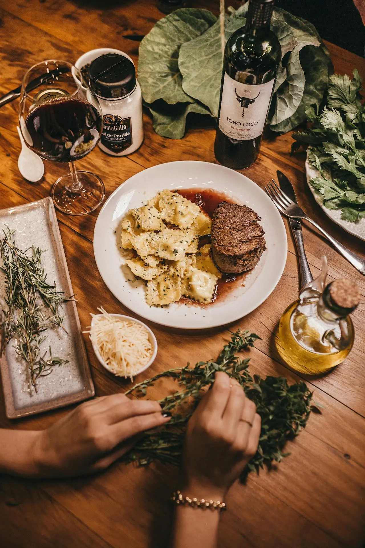 A plate with steak and ravioli sits on a wooden table surrounded by fresh herbs, a glass of red wine, a bottle of wine, grated cheese, olive oil, and other ingredients; hands arrange herbs in the foreground—perfect for those who visit Lyon.
