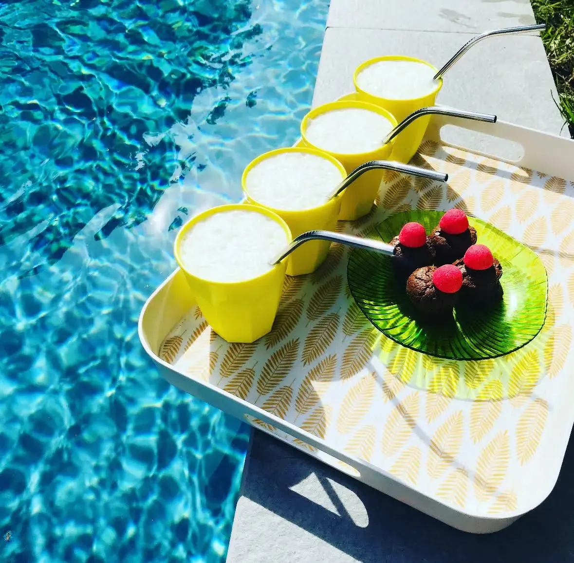 A tray by a swimming pool holds four yellow cups with metal straws and a green plate with three chocolate desserts topped with raspberries. Sunlight reflects on the water and tray, creating a bright summer mood.
