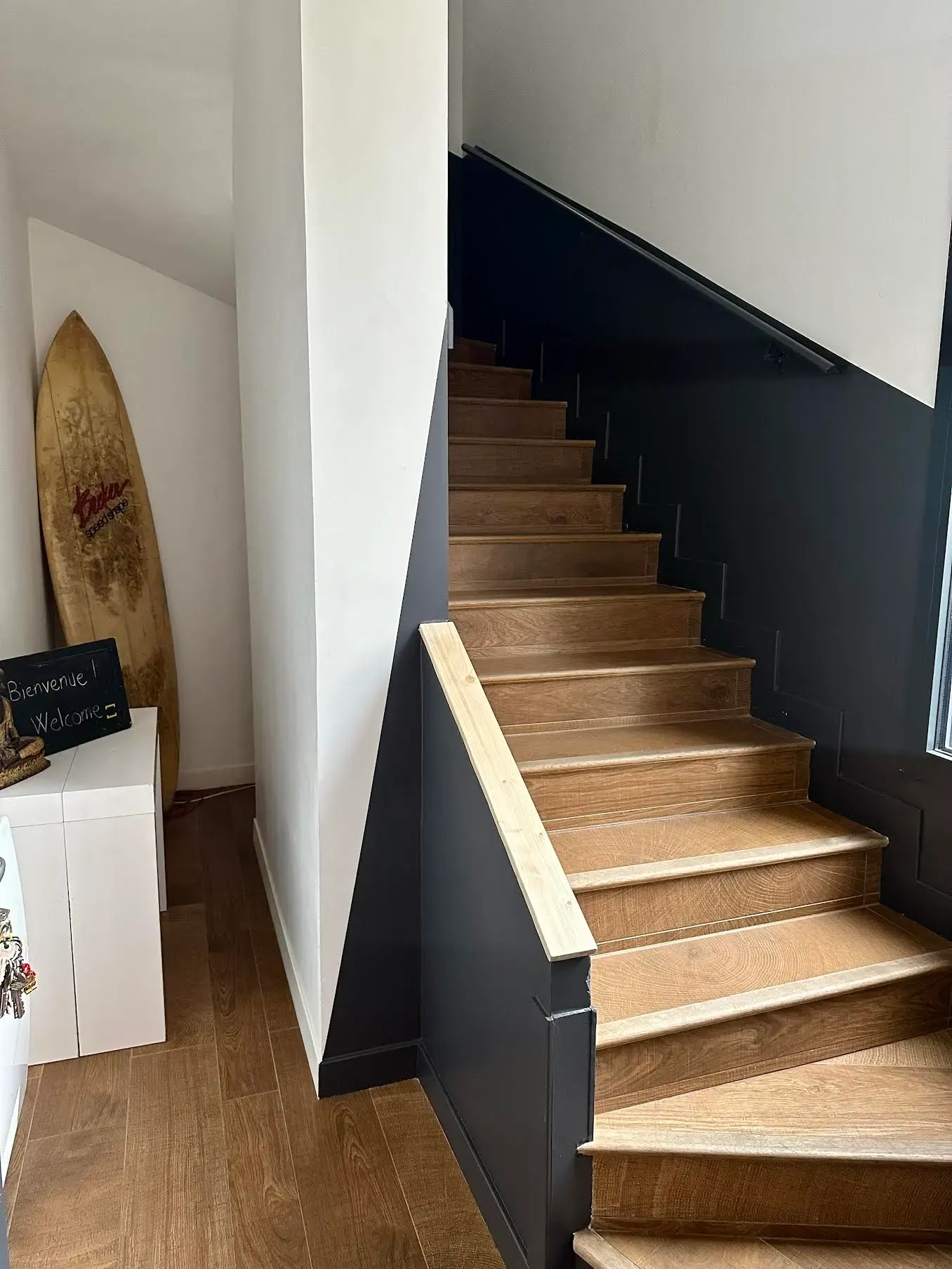 A modern wooden staircase with dark gray side panels is beside a small corner displaying a surfboard and a small white cabinet with a chalkboard sign that reads Bienvenue & Welcome.