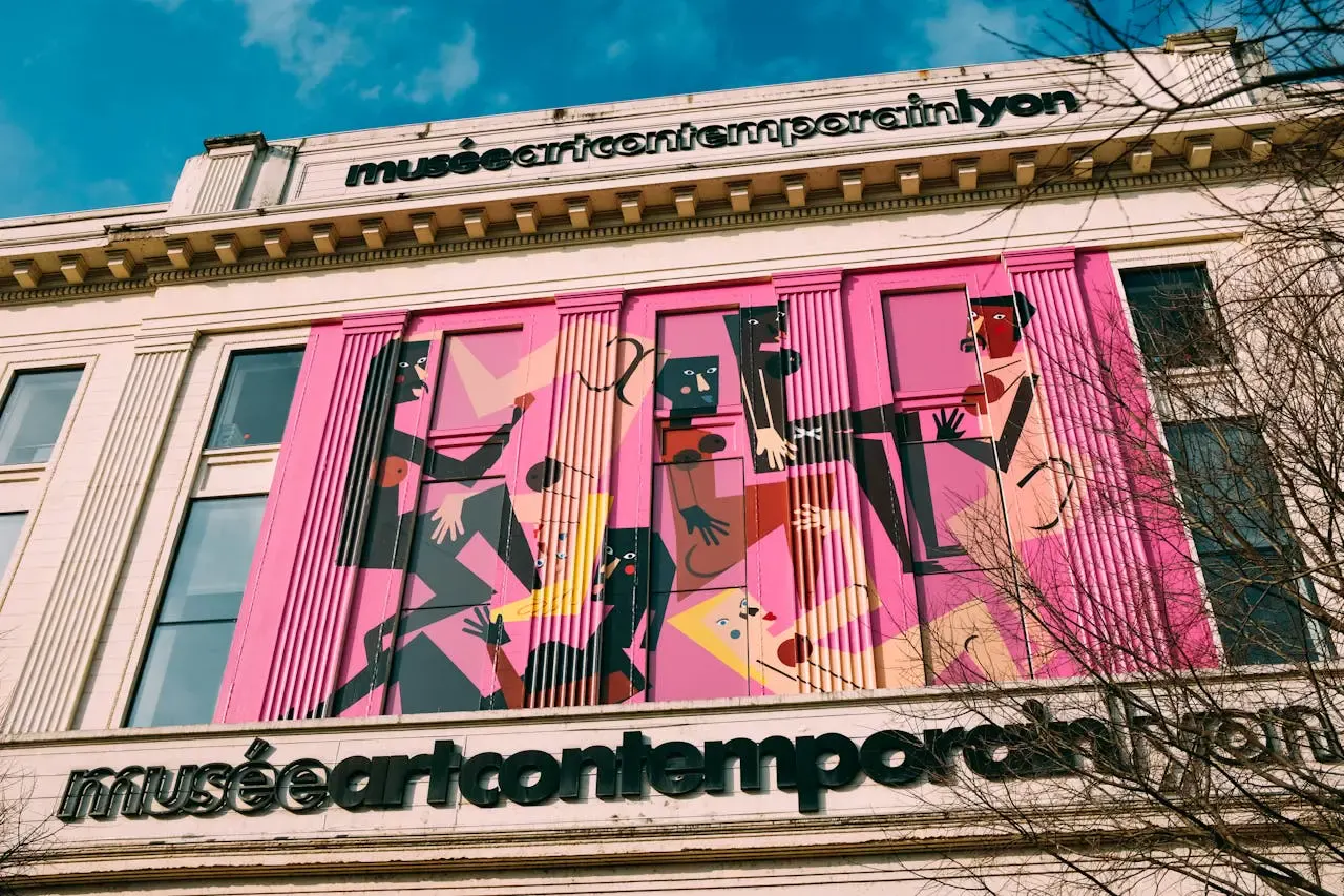 The exterior of the Musée d'art contemporain de Lyon features a vibrant mural with abstract, geometric human figures in shades of pink, purple, and yellow. Black text displaying the museum’s name stands out—an inviting sight for those who visit Lyon.