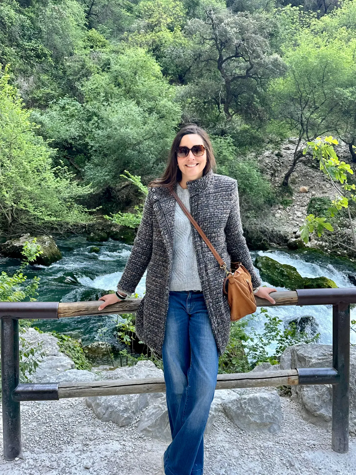 A woman in sunglasses, a gray coat, and jeans stands smiling in front of a wooden railing with a river and lush green trees in the background.