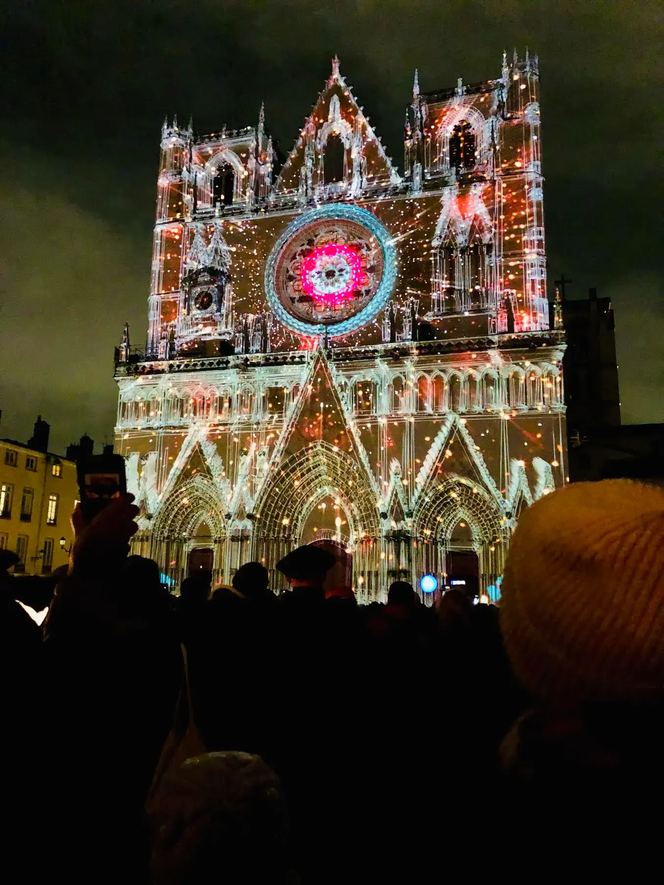 A cathedral illuminated at night with colorful light projections, including a bright circular design above the entrance, while a crowd of people watches in the foreground.