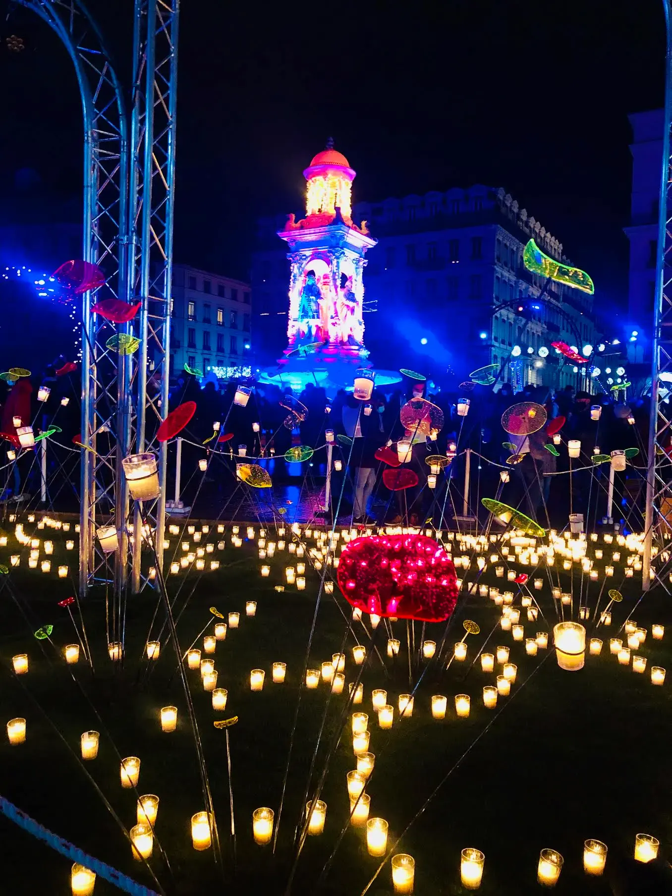 Scène de festival nocturne avec d'innombrables petites bougies disposées sur le sol, des décorations lumineuses colorées et une tour brillamment éclairée en arrière-plan, entourée de lumières bleues et roses incandescentes.