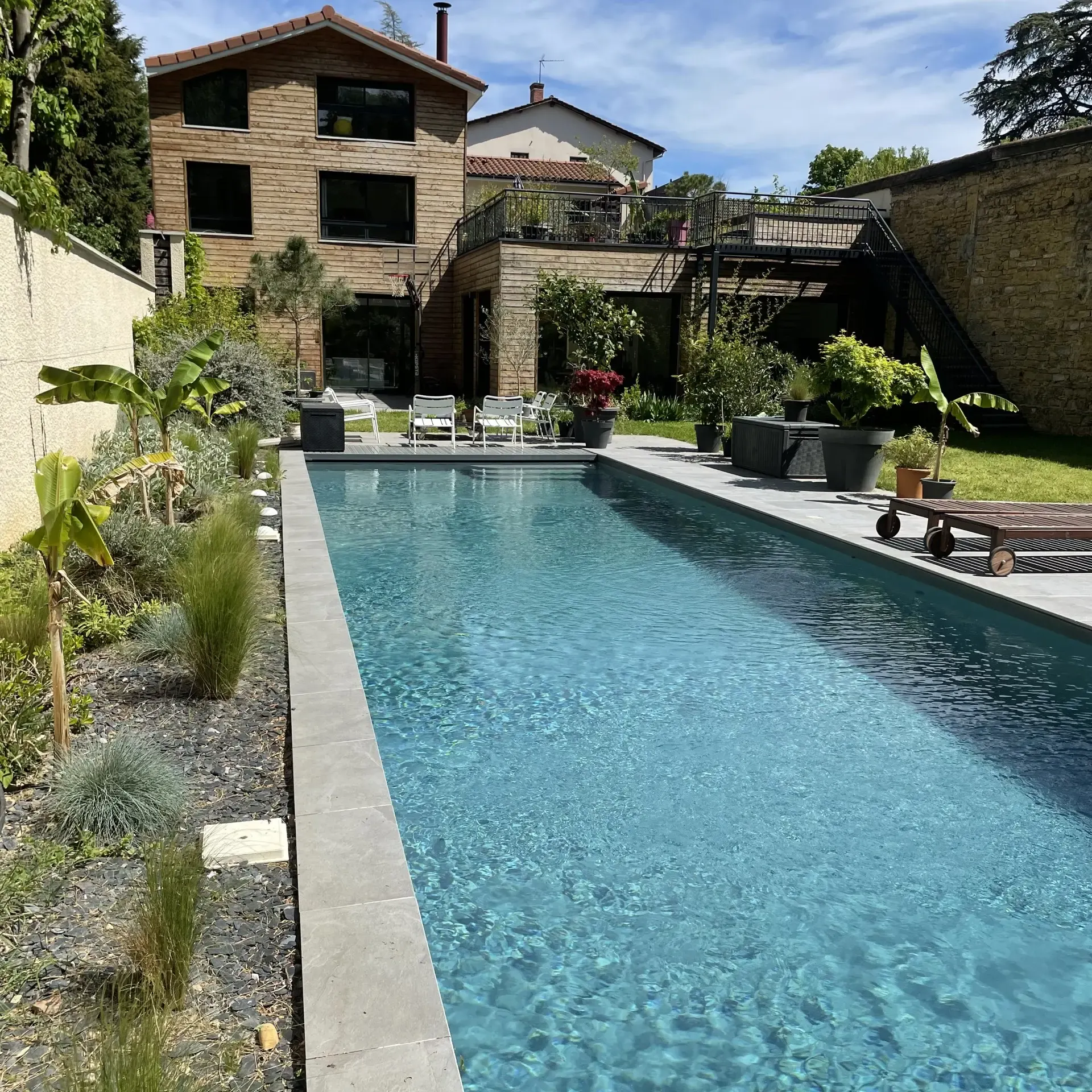Piscine de 15 mètres bordée de plantes tropicales, avec la maison en bois et pierre en arrière-plan