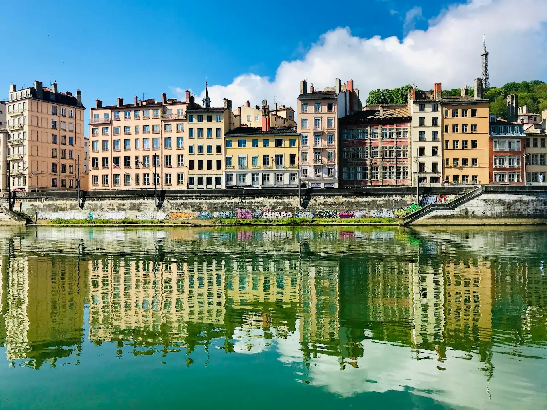 Des bâtiments colorés bordent une rivière, leurs reflets se reflétant dans l'eau calme et verdâtre en contrebas. Des graffitis décorent le mur du bord de la rivière et un ciel bleu vif avec des nuages cotonneux est visible au-dessus.