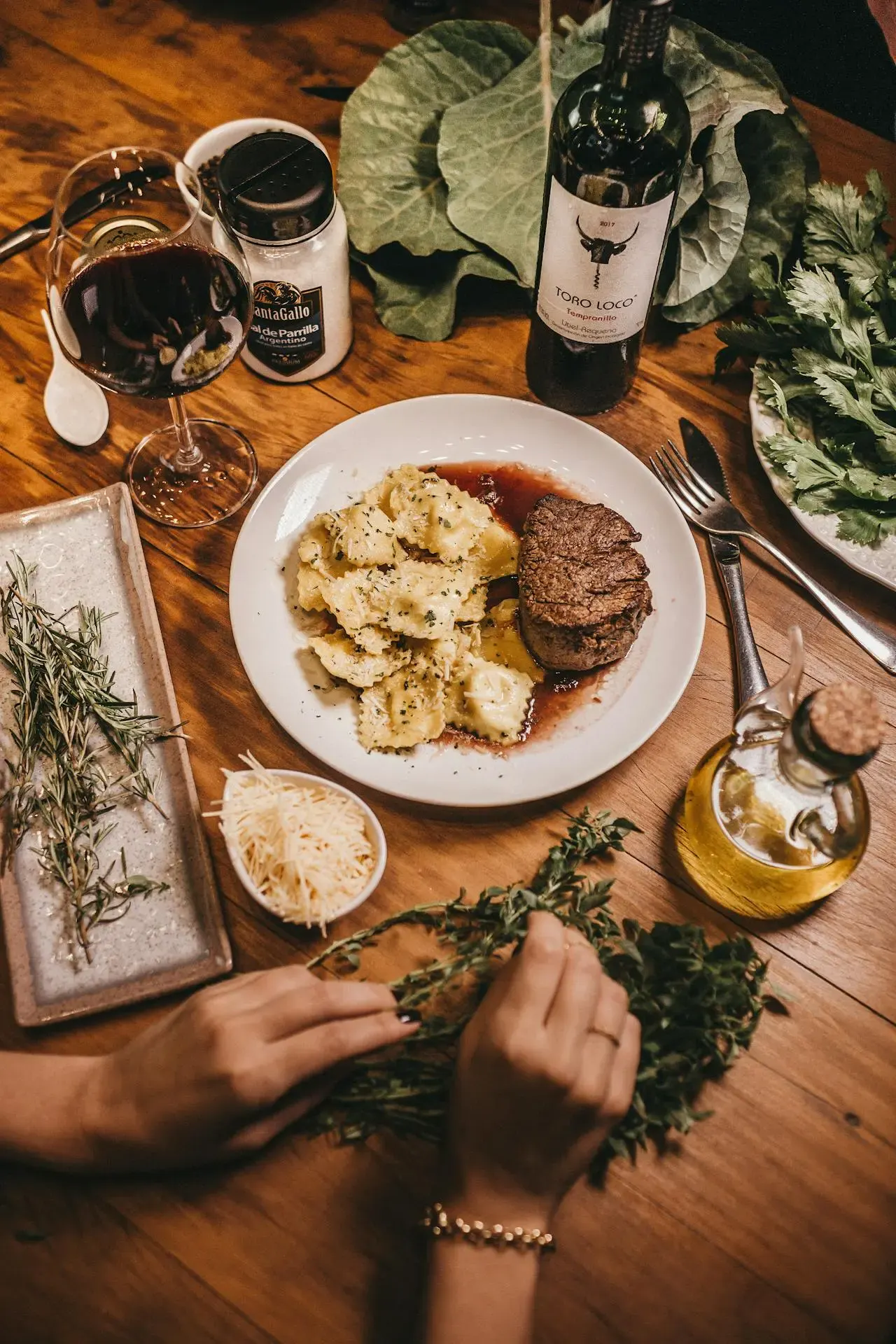 A plate with steak and ravioli sits on a wooden table surrounded by fresh herbs, a glass of red wine, a bottle of wine, grated cheese, olive oil, and other ingredients; hands arrange herbs in the foreground—perfect for those who visit Lyon.