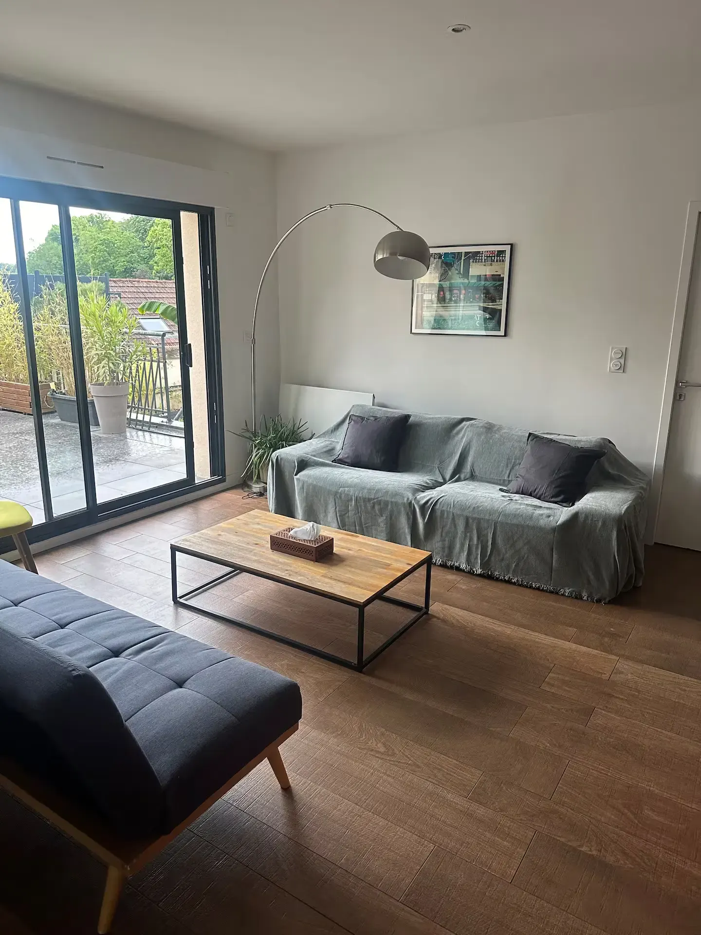 A modern living room in a guest house in Lyon features a gray sofa, dark futon, wooden coffee table, floor lamp, and framed picture. Large glass doors open to a balcony with plants outside.