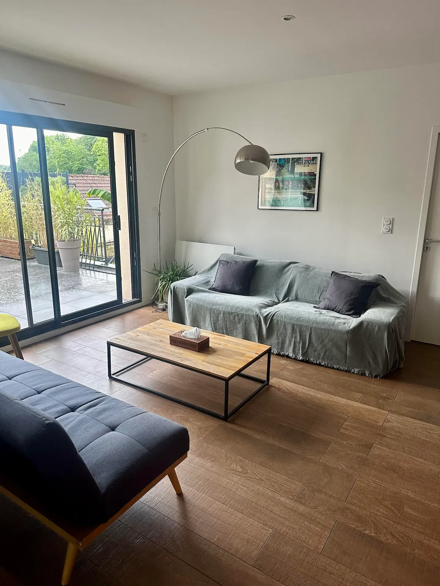 Un salon moderne avec des planchers en bois, un canapé gris, un futon noir, une table basse en bois, un lampadaire incurvé, un tableau encadré au mur et de grandes portes coulissantes en verre menant à un patio avec des plantes à l'extérieur.