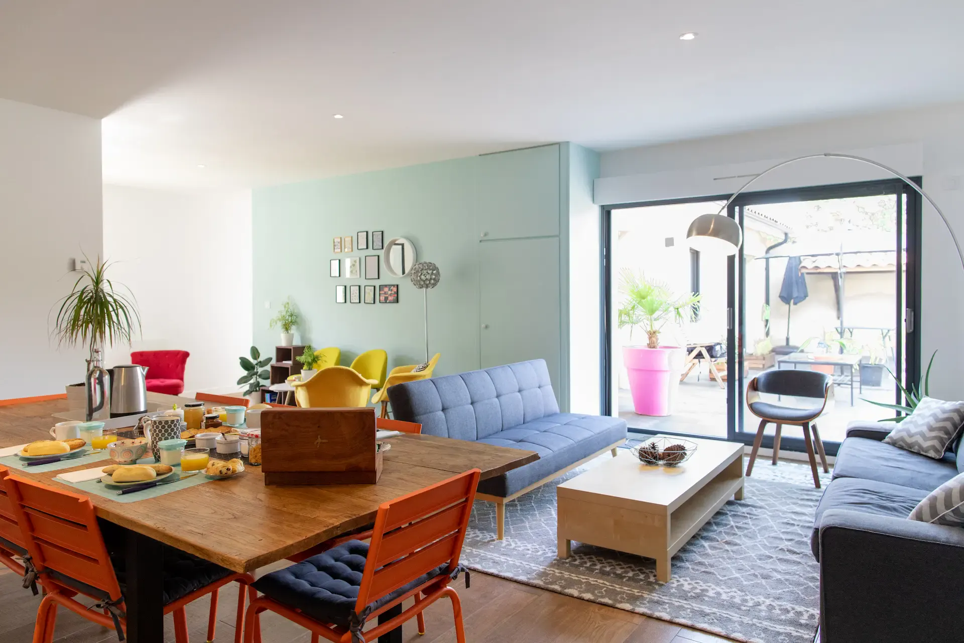 A bright, modern living and dining area in this guest house in Lyon features a wooden table set for breakfast, orange chairs, blue sofas, lush houseplants, and a gallery wall. Large glass doors open to a patio with plants and outdoor seating.