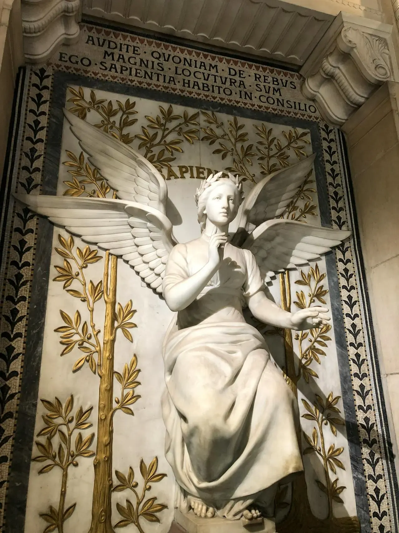A white marble statue of a seated angel with wings, in front of a decorated wall with gold laurel branches and a Latin inscription above. Visit Lyon to see the angel, who holds a finger to its lips, symbolizing silence or contemplation.