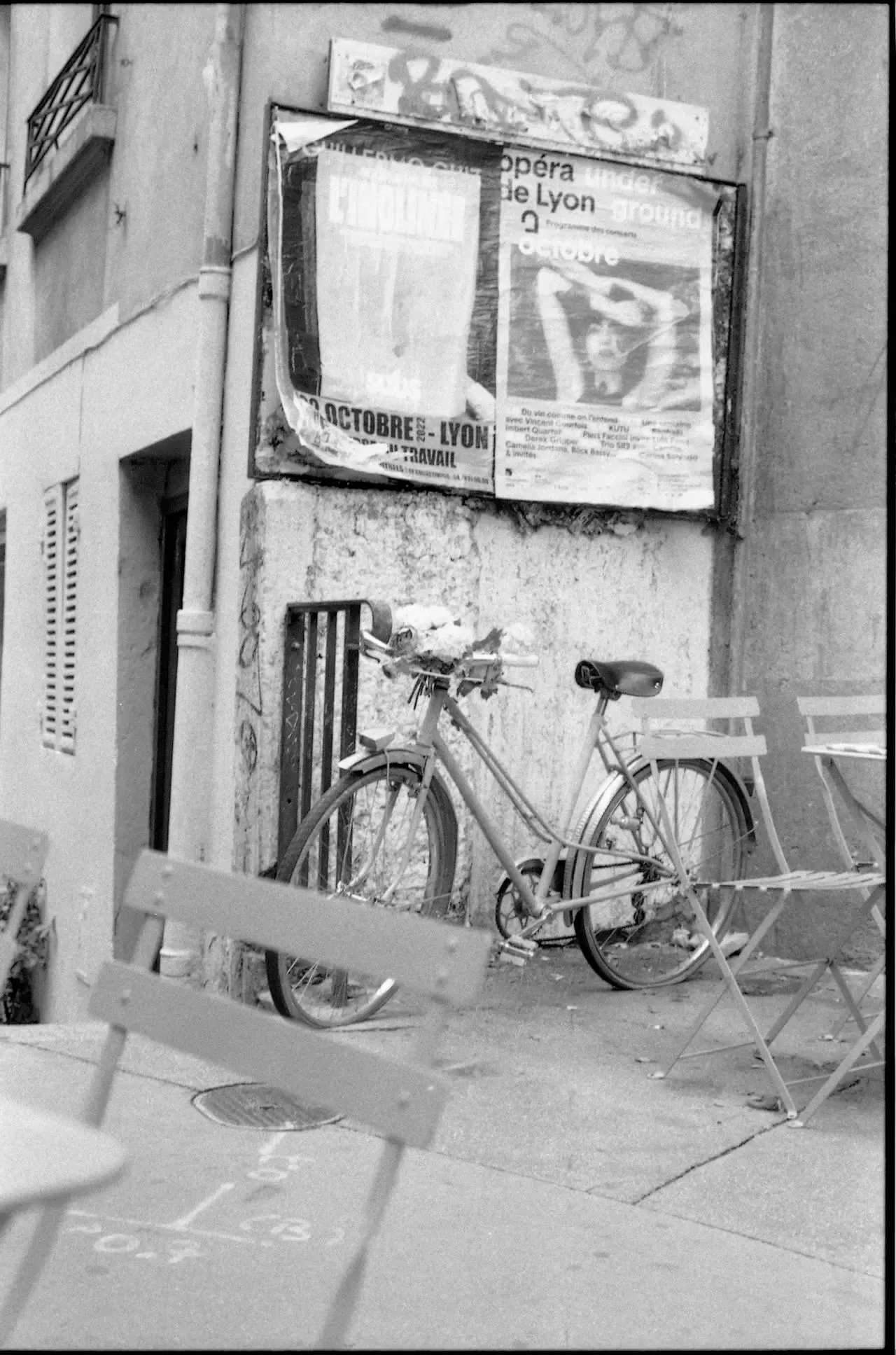 A black-and-white photo of a vintage bicycle leaning against a railing by weathered posters, near a building in Lyon. Folded café chairs and tables dot the slightly sloped street—capturing the charm that awaits when you visit Lyon.