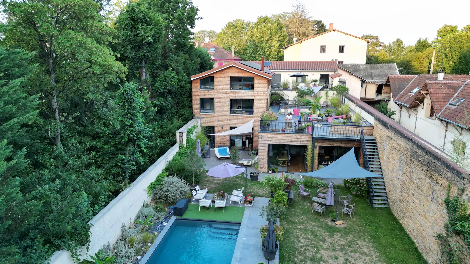Vue aérienne d'une maison moderne en bois avec de grandes fenêtres, entourée d'arbres. L'arrière-cour comprend une piscine, des meubles de terrasse, des zones ombragées avec des auvents et de la verdure. D'autres maisons sont visibles en arrière-plan.